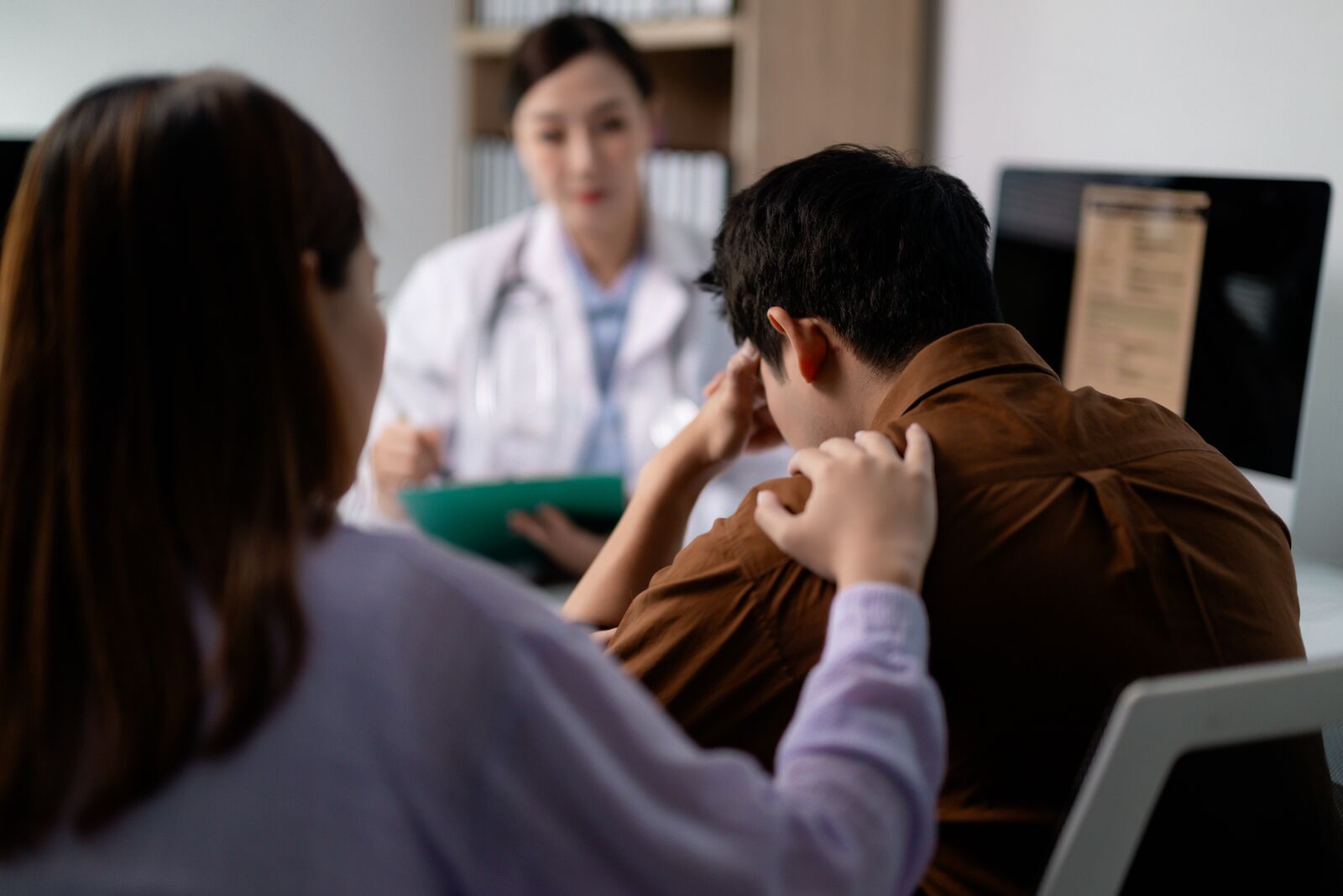 Woman resting a comforting hand on the shoulder of a man who lowers his head in distress while a doctor speaks with them across a desk. Woman resting a comforting hand on the shoulder of a man who lowers his head in distress while a doctor speaks with them across a desk.