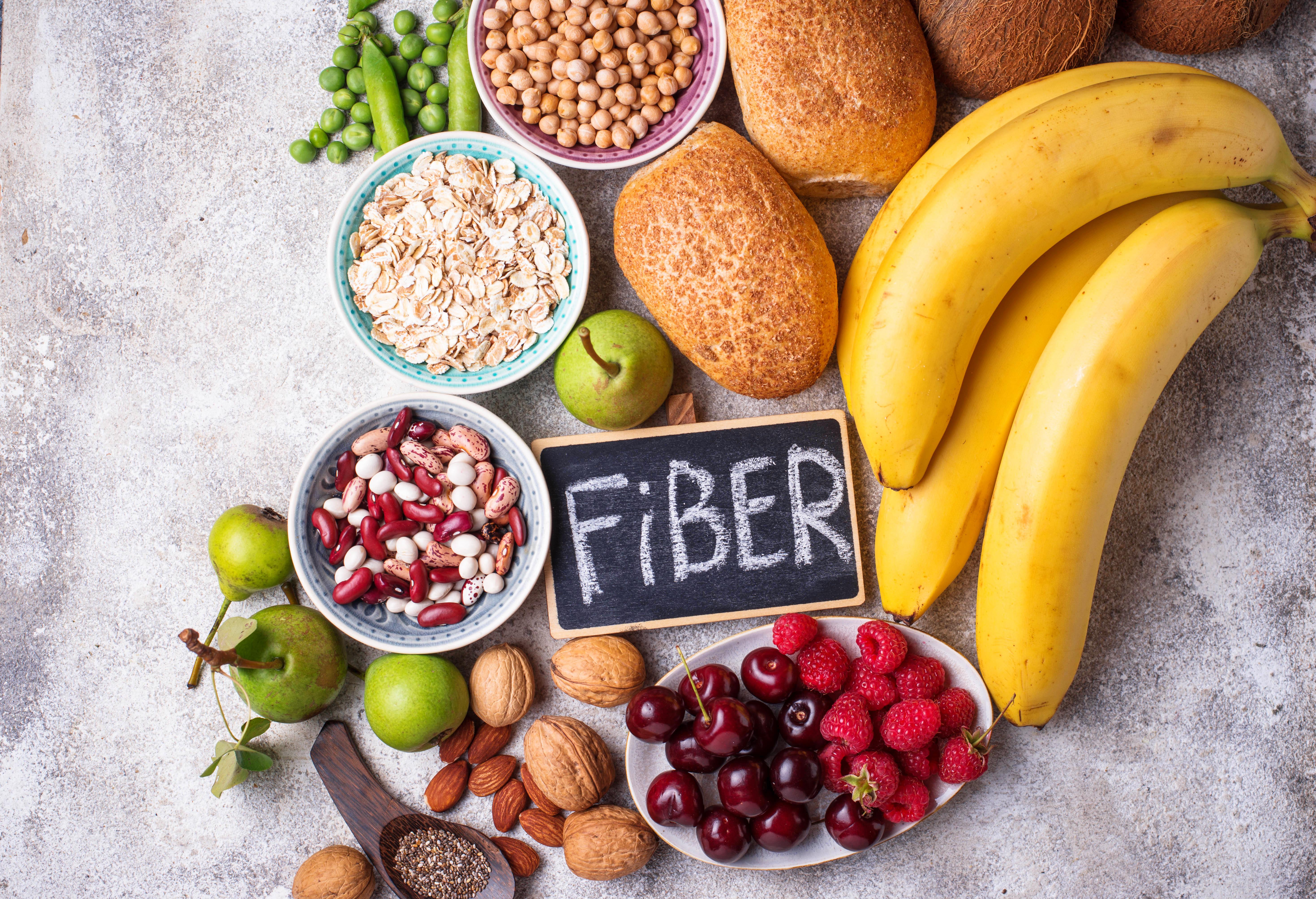 High-fiber foods displayed on a gray surface including bananas, berries, nuts, whole grain bread, legumes, oats, and fresh vegetables.