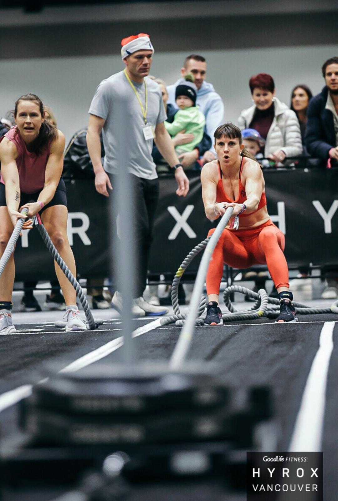 Two women perform battle rope exercises at a Goodlife Fitness Hyrox event in Vancouver with spectators watching. Two women perform battle rope exercises at a Goodlife Fitness Hyrox event in Vancouver with spectators watching.