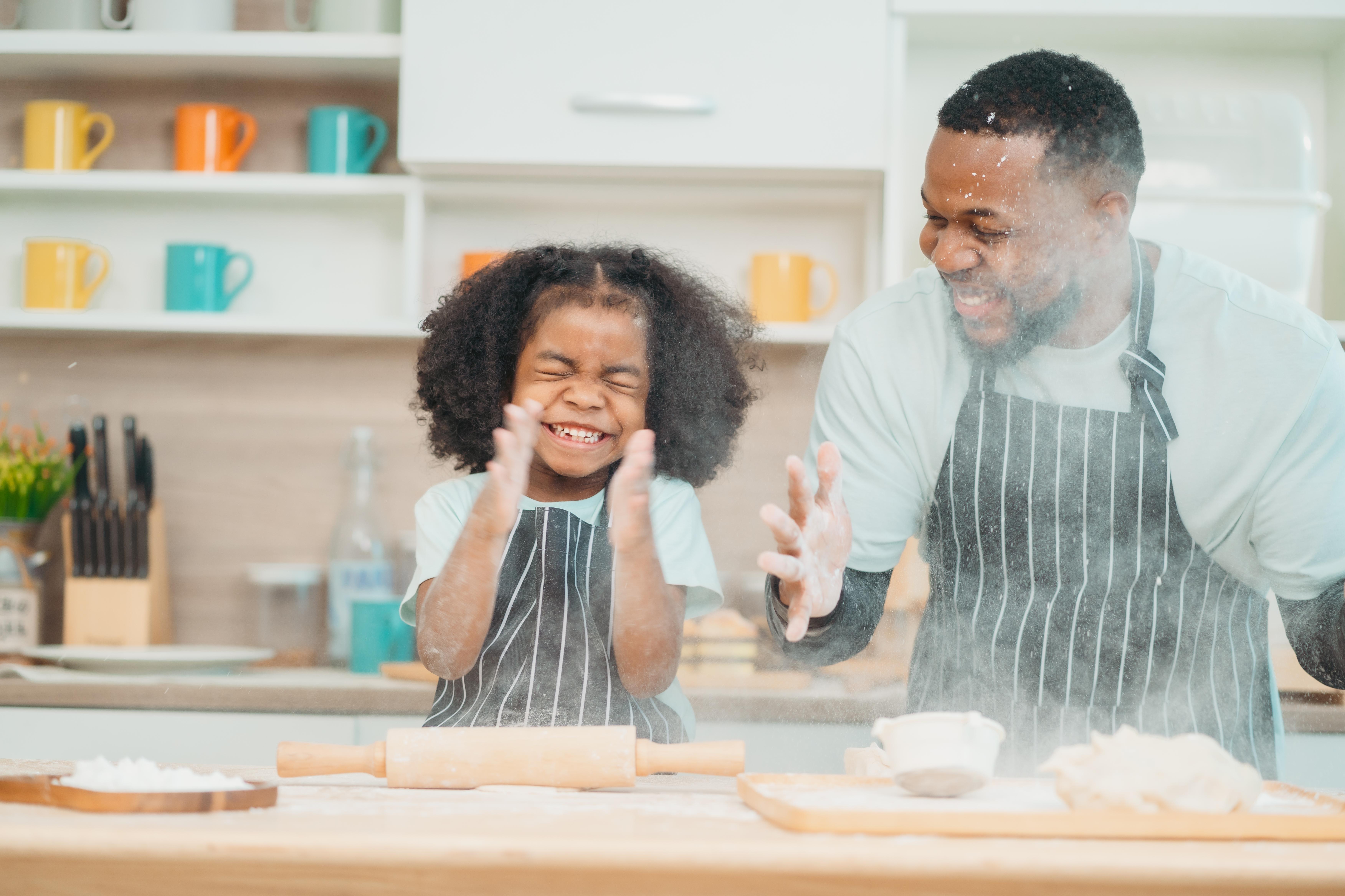 A father and young daughter wearing matching aprons bake together in a bright kitchen, both smiling as they work with flour and a rolling pin. A father and young daughter wearing matching aprons bake together in a bright kitchen, both smiling as they work with flour and a rolling pin.