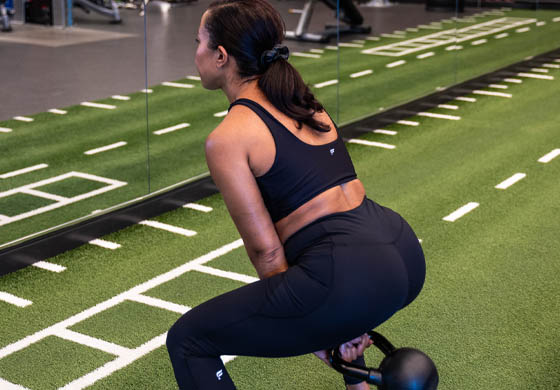 Young fit woman with her back to the camera doing a kettlebell swing on the turf, while facing the mirror, in the Toronto Athletic Club's Strength Lab