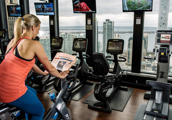 Blonde fit woman reading a newspaper on a stationary bike in the gym at the Toronto Athletic Club, view overlooking Lake Ontario and the city below
