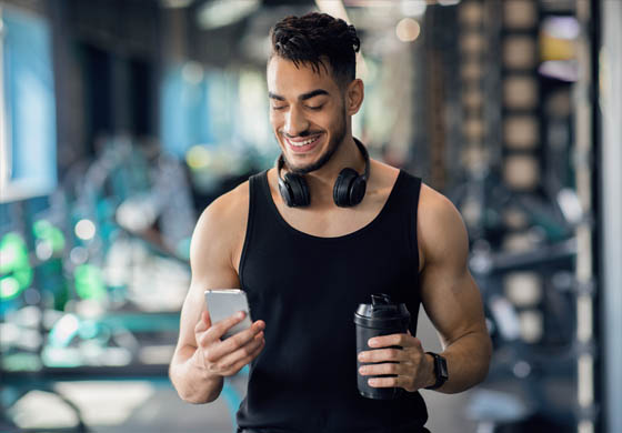 Young fit man in the gym with his protein shake looking at his phone, headphones around his neck