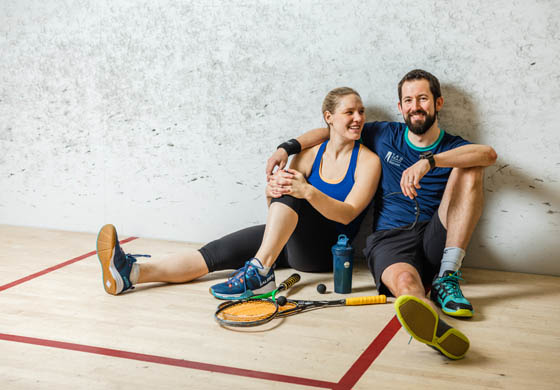 Man and woman sitting on the squash court, him with his arm over her shoulders and she's looking at him smiling; squash racuqets in front of them