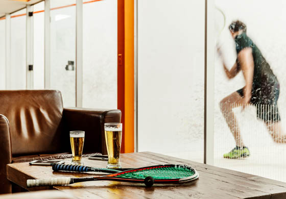 Young male member practicing on the squash court in the background, 2 pints on the table in the foreground