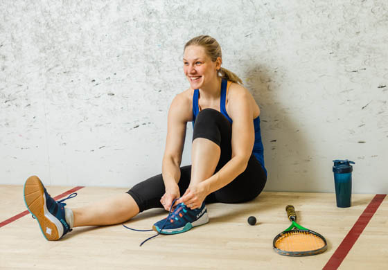 Young woman sitting on the squash court, tying her shoe, smiling at something off camera