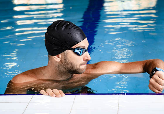 Young male with a swimming cap on, looking at his watch while he holds the edge of the pool